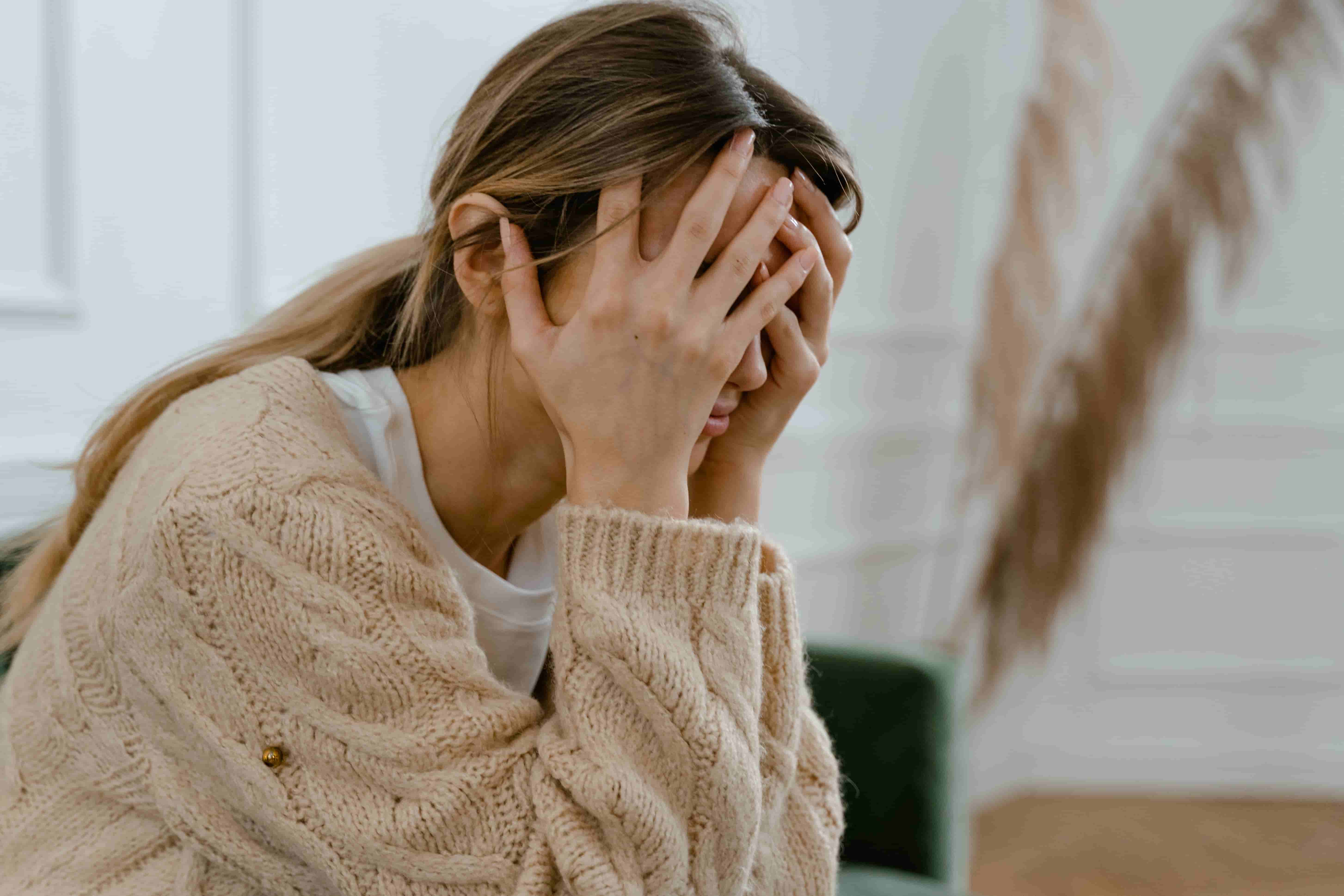 Junge Frau im Strickpullover verbirgt erschöpft das Gesicht hinter der Hand, Belastung nach einem einschneidenden Ereignis.