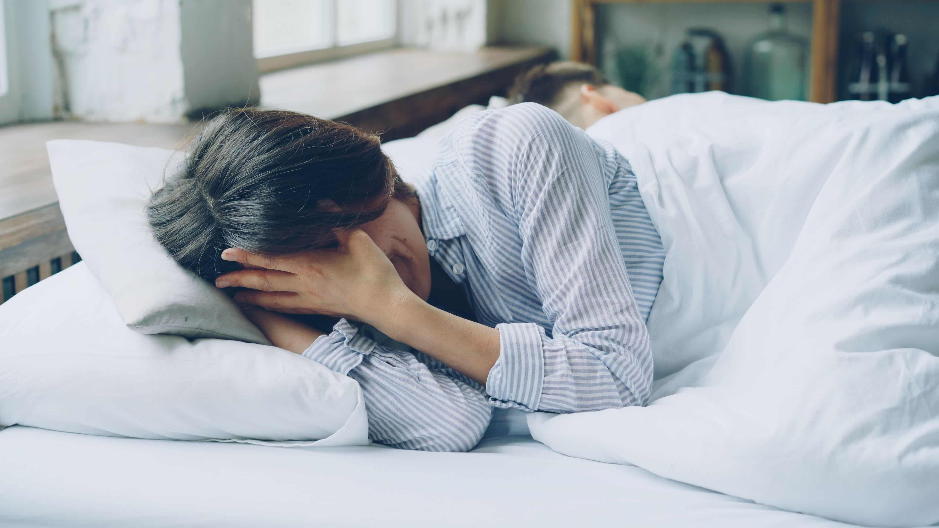 Frau liegt wach im Bett und hält sich den Kopf, neben ihr schläft jemand, Ein- und Durchschlafstörungen.